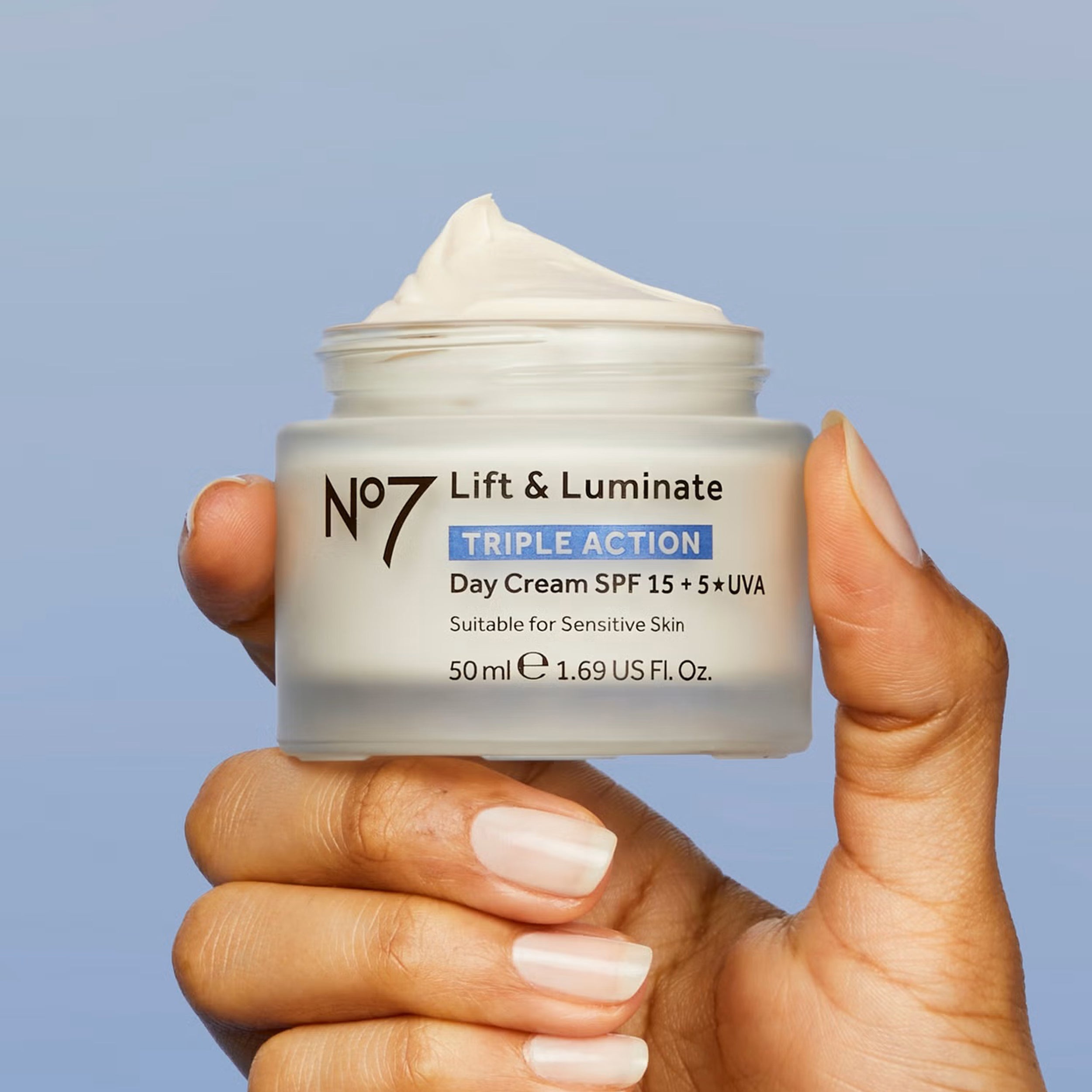 Hand holding a jar of No7 Lift & Luminate day cream against a blue background