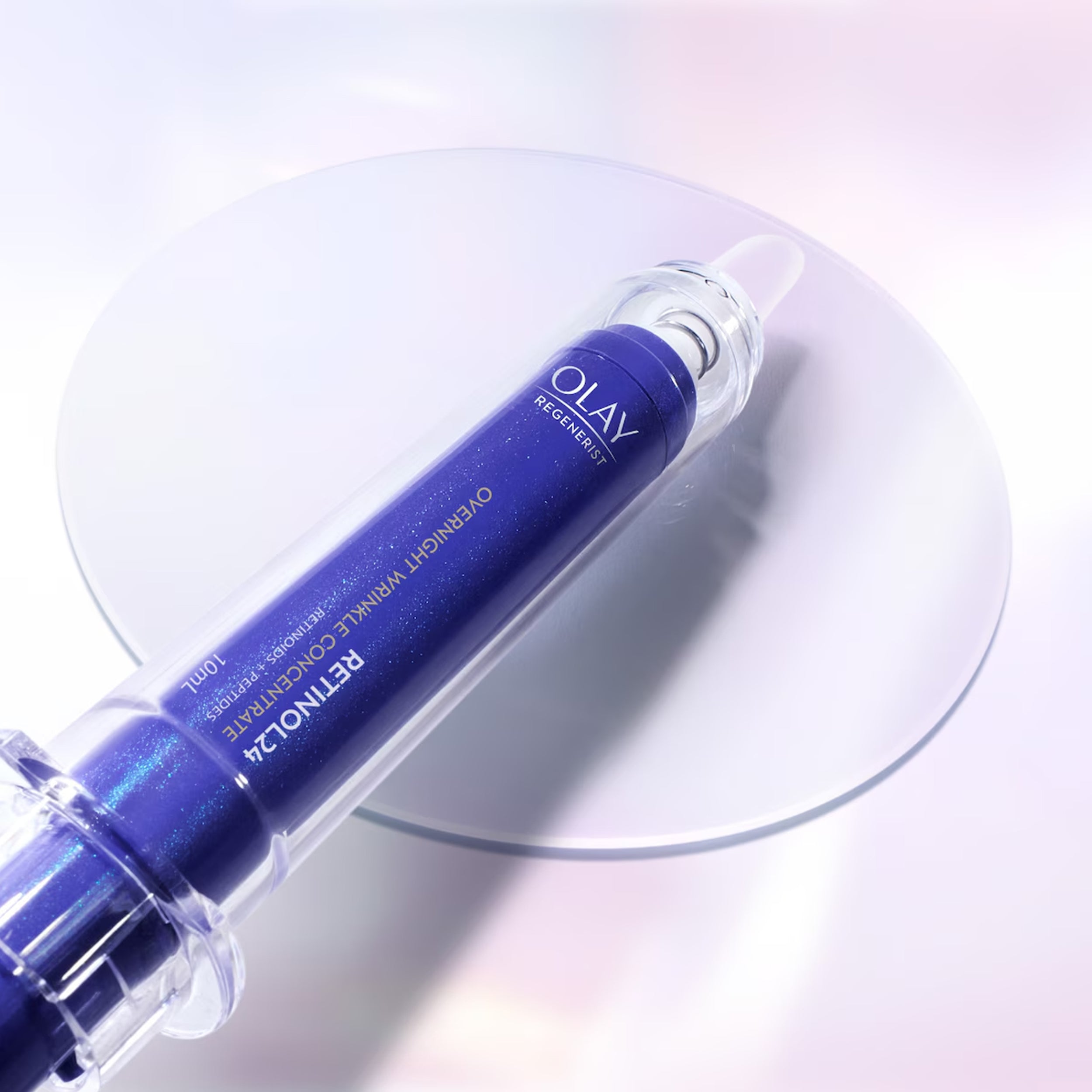 Olay Retinol 24% Overnight Wrinkle Concentrate in a deep blue cylindrical bottle with a clear cap, resting diagonally on a reflective circular glass plate against a soft white and pink gradient background.